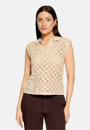 Femme portant un haut crochet beige sans manches avec col et un pantalon marron foncé sur un fond clair uni.