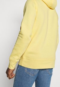 Sudadera amarilla de textura suave, con un bolsillo tipo canguro y puños acanalados. Combinada con jeans de mezclilla azules.