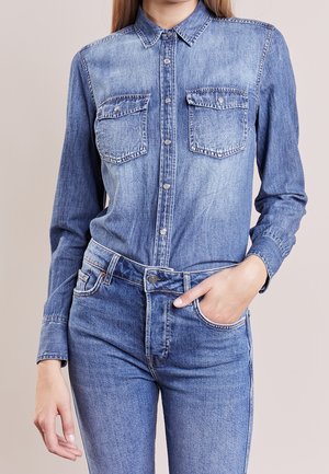 Kvinde iført lyseblå denimskjorte med knaplukning på klaplommer og matchende denimjeans, hånd i lommen, mod ensfarvet baggrund.