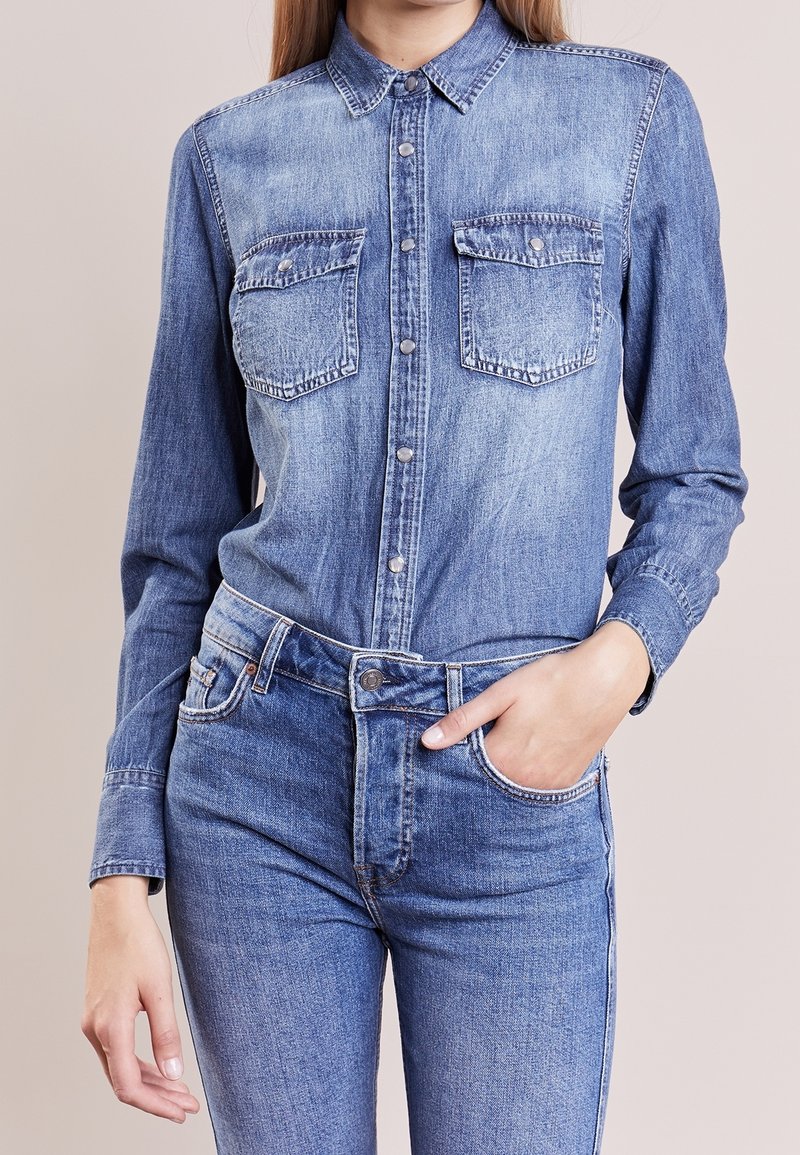 Femme portant une chemise en jean bleu clair avec poches à rabat boutonnées et un jean assorti, main dans la poche, sur fond uni.