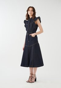 Dunkelblondes Denimkleid mit ausgestelltem Rock, strukturierten Schultern und kontrastierenden beigen Nähten. Mit Reißverschluss und seitlichen Taschen.