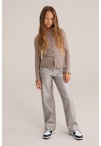 Junges Mädchen mit langen Haaren, das einen taubfarbenen Knopfcardigan, glänzende weite Hosen und schwarz-weiße Sneakers trägt und vor einem neutralen Hintergrund steht.