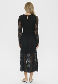 Juodas nėriniuotas suknelė su ilgu rankovėmis, raktų skylutės nugara ir laisvai krentančiu sijonu, išryškina sudėtingus nėrinių raštus ir elegantišką siluetą.