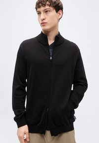 Μαύρο ζακέτα με φερμουάρ, κατασκευασμένο από μαλακό ύφασμα, με ψηλό γιακά, ριμπ μανσέτες και διακριτικό λογότυπο στο τελείωμα, με άνετη εφαρμογή.