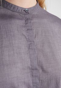 Camisa de lino morada con cuello redondo, con un pequeño cierre de botón y una textura sutil. Diseño simple con un corte recto.