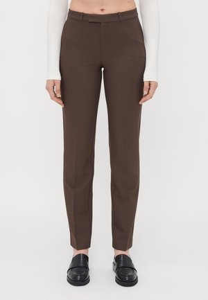 Schmal geschnittene braune Hose, kombiniert mit schwarzen Loafers und einem weißen langärmligen Top, das in der Taille gekürzt ist, vor einfachem Hintergrund.