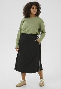 Jupe midi noire avec taille élastique et poches latérales, présentant une bande blanche contrastante. Associée à un haut à manches longues vert.