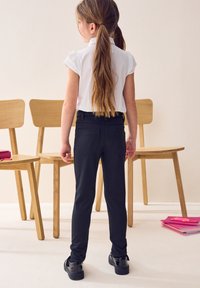 Pantalones ajustados negros con bolsillos traseros, combinados con una camisa blanca. El niño lleva zapatos negros. Al fondo, se pueden ver sillas de madera y útiles escolares.