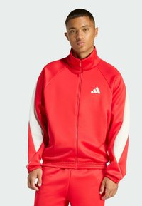 Red tracksuit made of smooth material with a high collar and white accents on the sleeves. Features a zip closure and an Adidas logo.