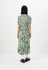 Grønn midilengde kjole med blomsterprint i svart og hvitt, korte ermer, elastisk midje og volangkant, sammen med kraftige svarte sko.