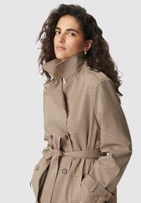 Brauner kariertes Trenchcoat mit hohem Kragen, Gürtel in der Taille und Knopfdetail. Besitzt eine weiche Textur und eine strukturierte Silhouette.