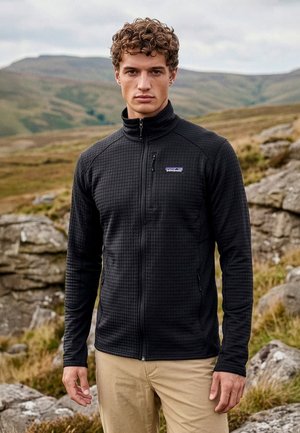 Nuori mies kiharat hiukset, musta vetoketjullinen takki ja beiget housut päällään, seisoo ulkona kalliolla, taustalla kukkuloita.