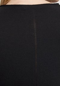 Gros plan du haut du dos d’une personne portant un vêtement noir texturé avec une couture verticale centrale, laissant apparaître la peau au-dessus de l’encolure.