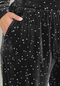 Svarta sammetsbyxor med spridda silverstjärnmönster, elastisk midja och fickor, som visar en mjuk textur och glans.
