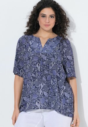 Vrouw met krullend haar, die een marineblauw topje met witte schelpmotieven en een witte broek draagt, staand tegen een effen achtergrond.