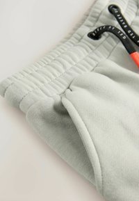 Helle graue Jogginghose aus strukturiertem Material, mit elastischem Bund und kontrastierendem orangenem Kordelzug. Verfügt über Taschen und Nähte als Akzente.