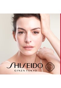 Nahaufnahme eines Frauengesichts mit braunen Augen und glatter Haut, die ein helles Oberteil trägt und deren Hände auf ihrem Kopf und ihrer Schulter ruhen.