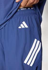 Μπλε αθλητικό σορτς κατασκευασμένο από ελαφρύ υλικό με πλεκτή μέση. Διαθέτει λευκό λογότυπο Adidas και τρεις διαγώνιες ρίγες στο πλάι.