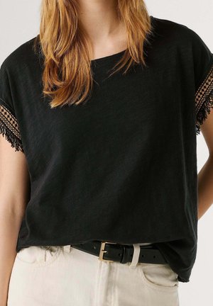 Woman wearing black short-sleeve top with lace trim on sleeves, beige pants, and a black belt with a silver buckle.