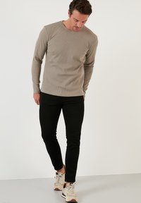 Long-sleeve beige shirt with a ribbed texture, paired with black slim-fit jeans and light-colored sneakers, standing on a plain background.