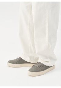 Baskets en daim gris à lacets avec semelles épaisses couleur écru portées avec un pantalon blanc ample sur fond blanc uni.