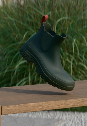 Bota de tobillo negra de goma con suela gruesa con dibujo y pestaña roja para tirar, situada encima de una superficie de madera con un fondo desenfocado de hierba verde.