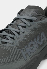 Grauer Laufschuh mit strukturiertem Mesh-Obermaterial, flachen Schnürsenkeln und auffälligem HOKA-Logo. Glatte Sohle mit dezenten Designmustern.