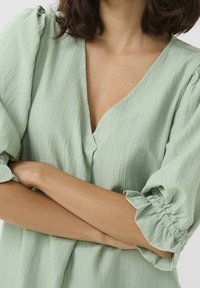 Blouse vert clair texturée avec un profond décolleté en V, manches bouffantes à fronces élastiques, et coupe ample. Boutons au centre devant.