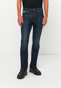 Muž v tmavomodrých slim-fit džínsach s jemným vyblednutím a čiernych kožených casual topánkach, stojaci pred jednofarebným bielym pozadím.