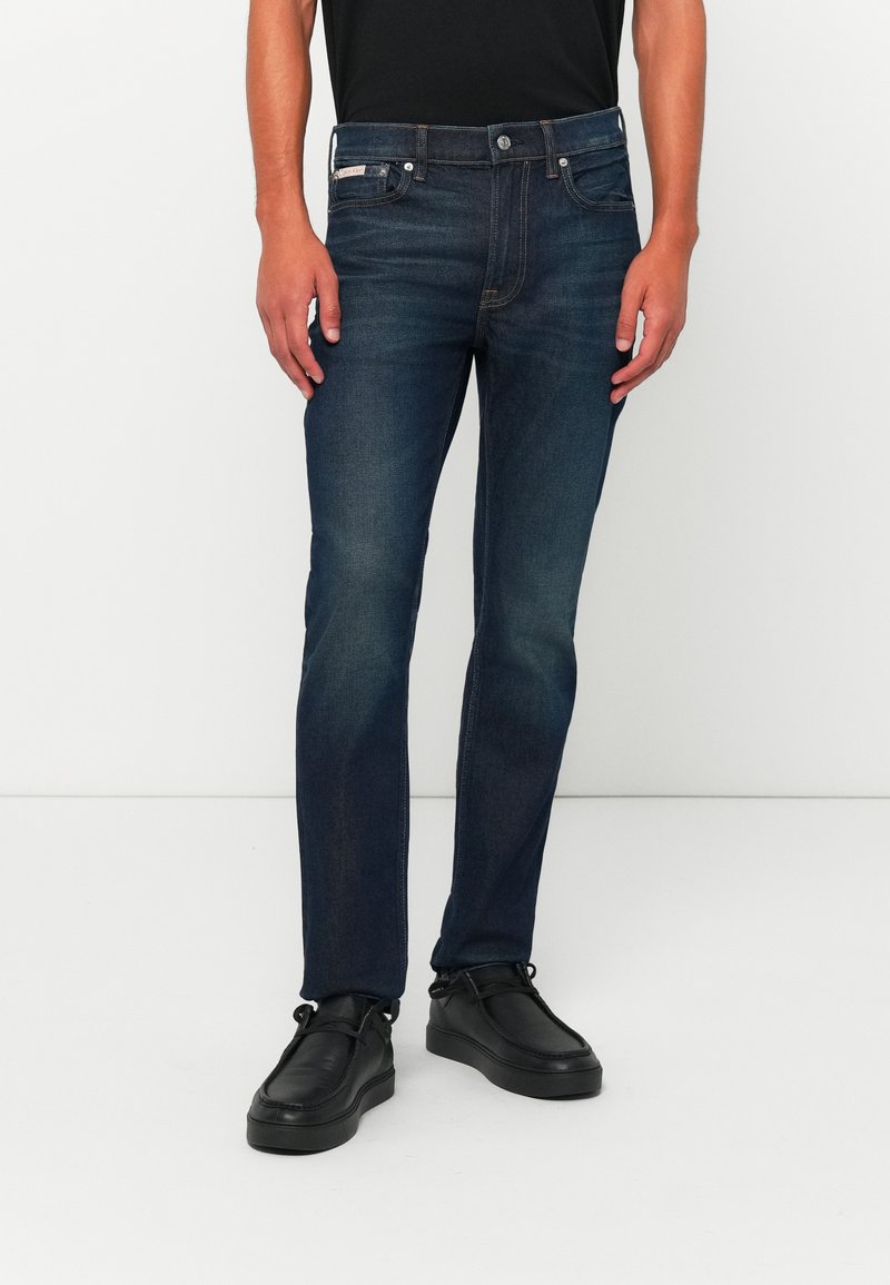 Muž v tmavomodrých slim-fit džínsach s jemným vyblednutím a čiernych kožených casual topánkach, stojaci pred jednofarebným bielym pozadím.