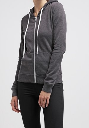 Femme portant un sweat à capuche zippé gris foncé avec des cordons blancs et un pantalon noir, debout devant un fond clair uni.