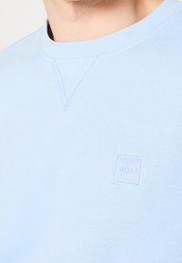 Sweat-shirt bleu clair à col rond avec un petit patch logo "BOSS" embossé sur la poitrine et un détail de couture triangulaire sous le col.