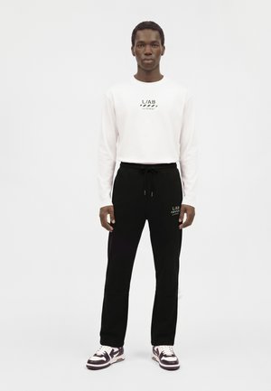 Mann trägt weißes Langarmshirt mit schwarzem Text und schwarze Kordelhose mit passendem Text, steht vor schlichtem weißem Hintergrund.