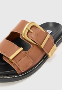 Sandale en cuir marron avec une grande boucle en or et une semelle noire. Présente des accents cousus et un design découpé sur la sangle supérieure.