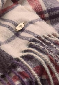 Skotsk uldhalstørklæde i nuancer af lilla, beige og rød; har frynsede kanter og et metalmærke med et detaljeret design.