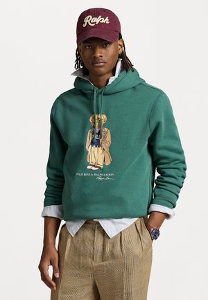 Groene hoodie van stof met een gestructureerde afwerking, voorzien van een kleurrijke beerafbeelding en gelaagd over een gestreept shirt, gecombineerd met geruite broeken.