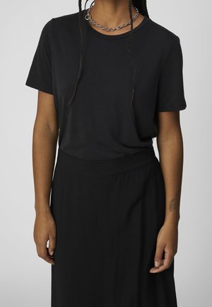 Person wearing a plain black short-sleeve shirt and matching skirt, with braided hair, a chain necklace, and a small floral tattoo on left forearm.