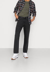 Jeans negros de corte recto, combinados con una camisa de franela a rayas en naranja, azul y blanco, superpuesta a una camiseta verde. Zapatillas blancas completan el conjunto.