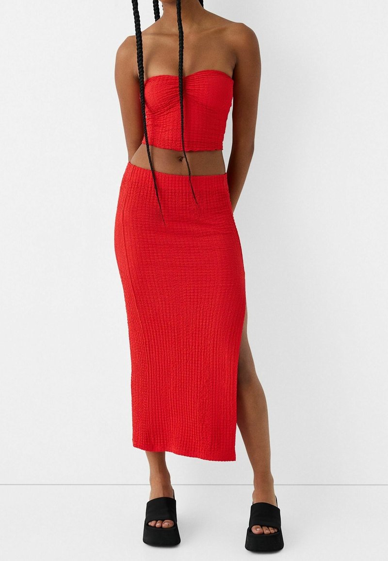 Ensemble rouge deux pièces composé d'un crop top bustier et d'une jupe mi-longue fendue sur le côté, fabriqués dans un tissu texturé. Sandales à talons carrés noires.