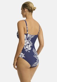 Kobieta stoi tyłem do kamery, ubrana w niebieski strój kąpielowy w kwiatowy wzór. Ma jasnobrązowe włosy upięte w kok.