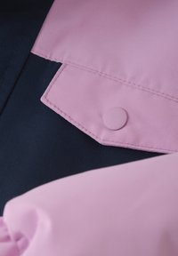 Två tyglappar, en mörk marinblå och en ljus rosa, med synliga sömmar och en rund rosa knapp. Texturerna är släta och matta.