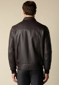 Muž viděný zezadu, oblečený v tmavě hnědé kožené bundě s žebrovanými manžetami a černých kalhotách na světlém béžovém pozadí.