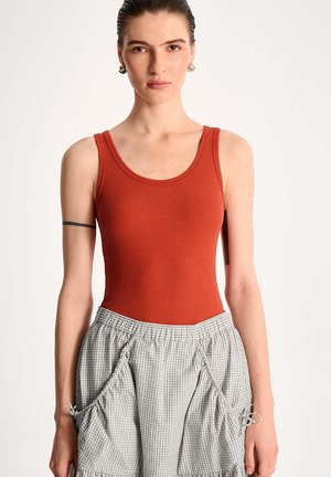 Kobieta w rudej, bezrękawnikowej bluzce i wysokim szarej kratce spódnicy z marszczonymi kieszeniami, stojąca na jednolitym tle.