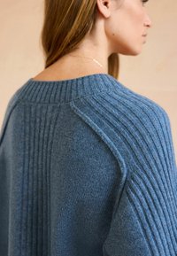 Blå strikket sweater med ribbet tekstur, bred halsudskæring og dropped shoulder-sømme. Stofmaterialet virker blødt og varmt.