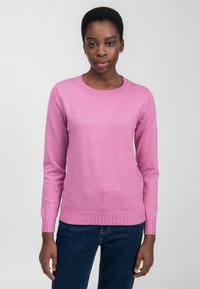 Donna con capelli corti che indossa un maglione rosa a maniche lunghe e jeans blu scuro su uno sfondo chiaro e uniforme.
