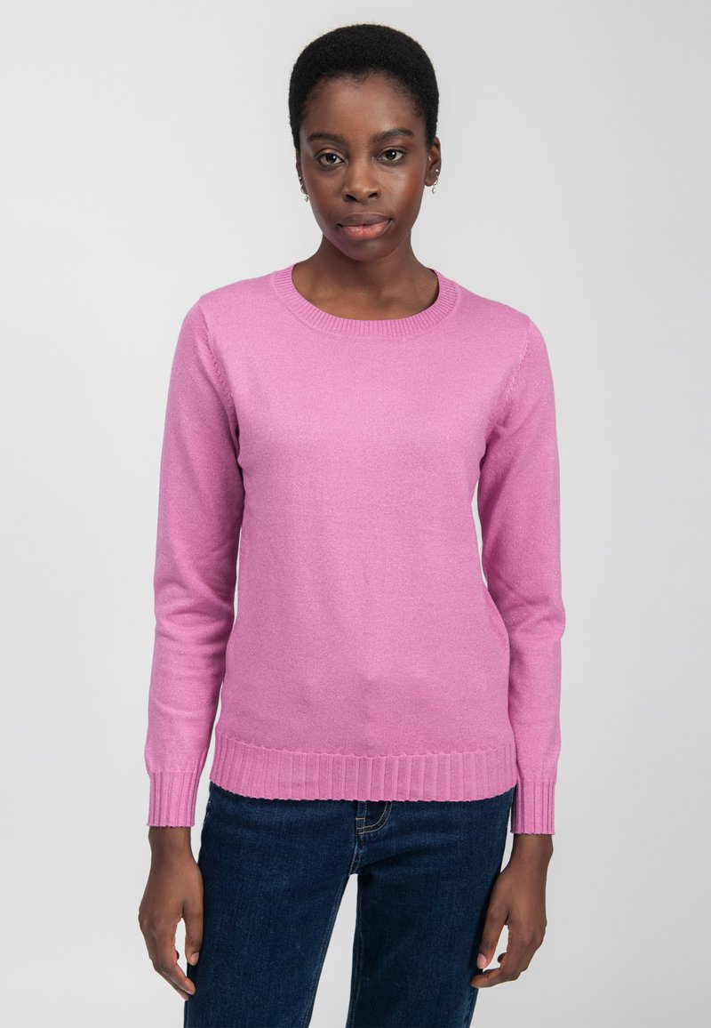 Donna con capelli corti che indossa un maglione rosa a maniche lunghe e jeans blu scuro su uno sfondo chiaro e uniforme.