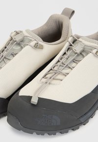 Двойка бежови и черни outdoor обувки The North Face с еластични връзки и текстурирана възглавничка на глезена, показани отблизо.