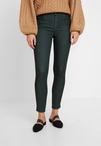 Jeans skinny verde oscuro con una textura suave y diseño tradicional de cinco bolsillos. Llevado con un suéter de punto trenzado color beige y zapatos negros de slip-on.