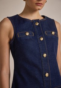 Top sans manches en denim bleu foncé avec des boutons décoratifs dorés, deux poches avant et des détails de surpiqûre contrastante.