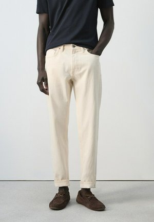 Hombre vistiendo pantalones de color crema remangados, camiseta negra de manga corta y mocasines de ante marrón, de pie contra un fondo claro y liso.